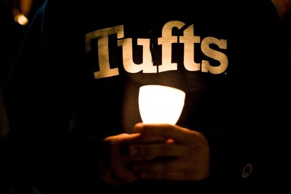 A person holds a small lit candle at their waist and it illuminates a Tufts logo on their sweatshirt; photo credit: Tufts University Photography